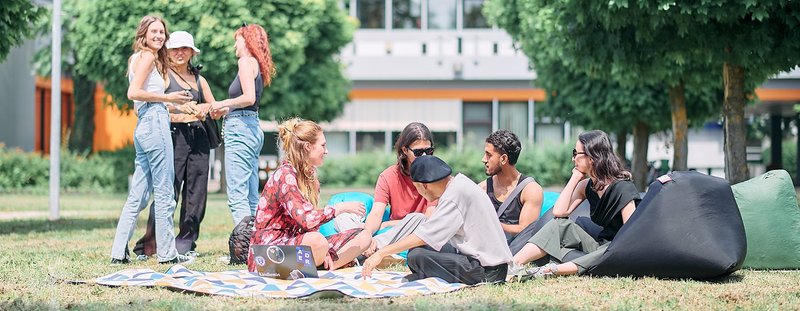 Studierende sitzen in der Sonne auf einer Wiese, unterhalten sich und spielen Ball