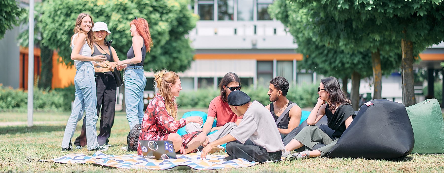 Studierende sitzen in der Sonne auf einer Wiese, unterhalten sich und spielen Ball